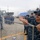 Sterett-Dewey Surface Action Group Deployment A group of military personnel in digital camouflage uniforms are gathered at a naval pier or port facility. A prominent sailor in the foreground wearing sunglasses and a navy cap marked '104' is handling a rope training rack, while a larger group of sailors stands in formation in the background. At least two naval vessels are visible docked in the background, including one marked '885'.