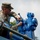 170615-N-YD204-030 A military service member in digital camouflage uniform is working on a large blue mechanical apparatus, likely a winch or hydraulic equipment. The individual appears to be performing maintenance or operational tasks, manipulating controls or fasteners on the blue machinery. The image is taken from a low angle, emphasizing the equipment and the focused work being performed.