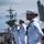 170614-N-BL637-029 U.S. Navy sailors in dress white uniforms are standing in formation on the flight deck of an aircraft carrier. The foreground sailor displays rank insignia, service ribbons, and a warfare qualification badge. A large aircraft carrier is visible in the background along with the tail section of a military jet aircraft.