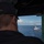 Bonhomme Richard Conducts Fueling at Sea with USS John S. McCain (DDG 56) A naval officer wearing a dark cap with 'CO' (Commanding Officer) insignia observes a warship from what appears to be a bridge wing or elevated vantage point of another vessel. The distant ship appears to be a U.S. Navy guided-missile destroyer underway at sea, visible through an opening framed by military equipment and wiring. The scene suggests an at-sea formation or replenishment operation under clear blue skies.