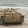 170614-N-PF515-007 A tracked armored fighting vehicle, consistent with a BMP-series infantry fighting vehicle, is positioned on a sandy beach with ocean waves visible in the background. The vehicle is painted in a tan/desert camouflage scheme and appears to be in motion or recently stopped, with sand disturbed around its tracks. A stack of white sandbags is visible to the right of the vehicle.