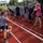Sterett-Dewey Surface Action Group Deployment A large group of people are participating in a tug-of-war competition on a red athletic track. Two distinct groups are visible — one wearing navy blue/white striped shirts and another in dark navy blue shirts — suggesting a friendly inter-group sporting event. Spectators line the sides of the track, cheering and laughing, indicating a festive, community-oriented atmosphere.