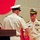 Commander, Amphibious Force 7th Fleet awards Capt. Paradise at Blue Ridge's change of command ceremony. Two U.S. Navy officers in white service dress uniforms are shaking hands at what appears to be a formal military ceremony, likely a change of command or award presentation. A wooden podium with a microphone is visible to the left, and a third officer is partially visible in the background. The officer facing the camera displays multiple service ribbons, gold warfare insignia, and a name tag.