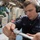 170614-N-UX013-408 A U.S. Navy sailor named Ward is performing maintenance or mechanical work aboard what appears to be a naval vessel. He is wearing green ear protection/hearing defenders and a dark navy blue coverall uniform while using a hand tool on a component. The background shows a cluttered shipboard workspace with pipes, equipment, and machinery.
