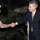 170613-N-FM530-037 A civilian in a dark suit and gold tie is shaking hands with a U.S. Marine Corps non-commissioned officer wearing tan service uniform and white dress cover (hat). The meeting appears to take place inside a military hangar or vehicle storage facility, with military vehicles visible in the background. A third uniformed individual is partially visible on the left edge of the frame.
