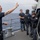 Sterett-Dewey Surface Action Group Deployment Military personnel are conducting a tactical training exercise on the deck of a naval vessel at sea. A sailor identified by name tape as 'Sullivan' is directing personnel with hand signals while others in tactical gear practice clearing a doorway/hatch with blue training firearms. The scene depicts a joint or combined arms boarding/security training drill.