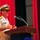 Commander, Amphibious Force 7th Fleet gives remarks at Blue Ridge's change of command ceremony. A U.S. Navy officer in white dress uniform is delivering a speech at a wooden podium with a microphone. The setting appears to be a formal military ceremony, indicated by the red curtain backdrop and the presence of at least one other uniformed officer in the background. The speaker's name tag is visible on his uniform, and he wears gold shoulder boards indicating senior officer rank.