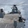 Sterett-Dewey Surface Action Group Deployment A military helicopter is hovering or landing on the flight deck of a naval warship at sea. The helicopter appears to be a large twin-rotor utility/transport type, positioned at the stern flight deck of the vessel. The ship is underway in open blue ocean waters under clear daylight conditions.