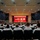 Commander, Amphibious Force 7th Fleet gives remarks at Blue Ridge's change of command ceremony. A large auditorium filled with military personnel dressed in white uniforms attending a formal ceremony or briefing. The stage at the front displays both the American flag and the Japanese flag against a red curtain backdrop, with several individuals seated or standing on stage. The setting strongly suggests a joint US-Japan military event, likely held at a US military installation in Japan.