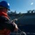 170614-N-XK398-079 A naval sailor wearing a blue hard hat and orange life vest is handling lines or cables during what appears to be an underway replenishment (UNREP) or ship-to-ship transfer operation at sea. A large naval vessel, likely an amphibious assault ship or aircraft carrier, is visible in close proximity alongside. The operation is taking place in open ocean under clear blue skies.
