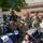 NMCSD NEC Fair Two U.S. Navy sailors in camouflage uniforms are speaking with a civilian woman at an outdoor military career or recruiting fair. The display booth features deep sea diving equipment and informational boards about Navy diving specialties, including the '8494 Deep Sea Diving Independent Duty Corpsman' and '8493 Deep Sea Diving Medical Technician' ratings. The event appears to be held in a courtyard of a military or government facility.