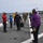 170614-N-JI086-354 The image shows a military flight deck ceremony or arrival protocol aboard what appears to be a naval amphibious assault ship. Personnel in color-coded flight deck jerseys (purple, red, blue, yellow) are lined up on either side of the deck, forming a corridor as officers walk through. A V-22 Osprey tiltrotor aircraft is visible on the deck in the background.