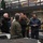 170614-N-JI086-354 A multinational military briefing is taking place inside what appears to be a naval vessel's hangar deck or cargo bay. Personnel in various national military uniforms are gathered around a US Navy officer in a flight suit who appears to be conducting a tour or briefing. The group includes representatives wearing German Bundeswehr, Polish, Italian, and US Navy uniforms.