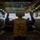 170614-N-GG458-004 A naval crew member is seen from behind, standing in the well deck of a U.S. Navy amphibious transport dock ship, holding orange marshalling wands above their head. The person is wearing a yellow safety vest marked '05' and 'LPD-24', along with a yellow helmet and hearing protection. The well deck appears to be open at the stern, with a landing craft or vehicle visible in the background.