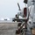 170602-N-FM530-135 A military attack helicopter, consistent with a Bell AH-1Z Viper, is chained down on the flight deck of a naval amphibious assault ship. In the background, a Landing Craft Air Cushion (LCAC) hovercraft is visible approaching or departing the vessel across open ocean. The scene depicts a multi-asset amphibious naval operation.