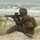 170614-N-PF515-008 A military service member in MARPAT (Marine pattern) digital camouflage uniform is positioned on a sandy beach dune in a kneeling firing stance. The soldier is aiming an M16/M4-style rifle equipped with an optical scope toward an unseen target. The setting appears to be a coastal training exercise, indicated by the beach environment and a red training/blank firing adapter on the rifle muzzle.