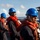 170614-N-XK398-036 A group of naval personnel are standing in formation on the deck of a military vessel at sea. All individuals are wearing blue hard hats and orange life vests over dark navy uniforms, suggesting a safety drill or deck evolution exercise. The setting appears to be an active warship with superstructure visible in the background.