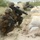 170614-N-PF515-009 The image shows what appears to be U.S. Marines in full combat gear conducting a military training exercise on a sandy beach environment. Three personnel in MARPAT (Marine Pattern) camouflage uniforms are positioned behind a row of sandbags in tactical crouching stances. The soldiers are armed with assault rifles and appear to be practicing defensive or assault maneuvers.