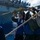 170614-N-XK398-032 Naval personnel are conducting a replenishment-at-sea (RAS) or underway replenishment (UNREP) operation aboard a military vessel. Sailors wearing blue coveralls, hard hats, and work gloves are handling heavy mooring or transfer lines. A large warship, likely an aircraft carrier or amphibious assault ship, is visible in the background alongside.