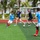 Sterett-Dewey Surface Action Group Deployment A soccer match is being played on an artificial turf field between two teams — one wearing light blue jerseys with white shorts and red socks, and the other wearing dark navy shirts. The game appears to be an informal or friendly match, possibly between local players and foreign visitors, set in what appears to be a coastal Asian city based on the urban skyline and tropical vegetation visible in the background. The action is centered around a contested ball near the middle of the field.