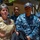 NMCSD NEC Fair A group of U.S. Navy personnel are gathered outdoors, with a senior female officer in tan/khaki service uniform appearing to address or brief the group. Several enlisted sailors in Navy Working Uniform (NWU) digital camouflage pattern are listening attentively. The setting appears to be an urban or base environment near a building with an elevators sign visible.