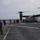 170614-N-JI086-354 A military tiltrotor aircraft, identifiable as a V-22 Osprey, is parked on the flight deck of a naval vessel at sea with its rotors spinning. Multiple personnel in mixed military uniforms and civilian attire are disembarking or moving away from the aircraft. A flight deck crew member in a purple vest and orange helmet stands in the foreground, likely serving as a handler or safety observer.