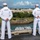 170614-N-BL637-020 Two U.S. Navy sailors in dress white uniforms stand at parade rest on the deck of a naval vessel, overlooking a waterway near Hickam Air Force Base in Hawaii. A crowd of people has gathered along the shoreline to watch the ship's arrival or departure. The cityscape of Honolulu and lush tropical vegetation are visible in the background.