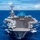 170612-N-BL637-093 A large US Navy aircraft carrier is underway at sea, viewed from a forward elevated angle showing the full flight deck. The carrier is loaded with numerous fixed-wing aircraft and has personnel visible on deck. A second naval vessel is visible in the background at distance.