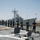 170614-N-FQ994-169 Naval personnel in dark blue uniforms are standing at attention in a line along the deck of a warship, facing a pier where another naval vessel is docked. The scene appears to depict a formal naval ceremony, likely a 'manning the rails' or port arrival/departure procedure. A flag, possibly Spanish, is visible on a flagpole in the background near port facilities.