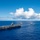 170612-N-BL637-038 A large naval carrier strike group is underway in open ocean under clear blue skies with scattered cumulus clouds. The formation is led by a Nimitz-class or Gerald R. Ford-class aircraft carrier with aircraft visible on its flight deck, accompanied by three escort vessels. A fixed-wing aircraft is also visible flying low to the left of the carrier.