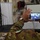 170614-N-JI086-354 A military personnel wearing Italian Army camouflage uniform is recording a ceremony or briefing on a smartphone. The phone screen displays a 'Welcome Aboard' banner with what appears to be a naval insignia, suggesting the event is taking place aboard a naval vessel. In the background, a speaker stands at a podium flanked by flags in an interior ship compartment.