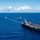 170612-N-BL637-055 An aerial photograph shows a naval carrier strike group underway in open blue ocean waters. A large aircraft carrier dominates the foreground with multiple fixed-wing aircraft visible on its flight deck, while four escort vessels — likely destroyers or cruisers — are arrayed in formation behind it. The formation is sailing under partly cloudy skies in what appears to be a clear, calm sea environment.
