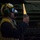 170614-N-GG458-030 A military deck crew member wearing a yellow safety vest and yellow helmet with ear protection holds an illuminated orange/yellow marshalling wand upright. A second crew member in a white helmet is visible in the background. The setting appears to be a dimly lit hangar bay or flight deck aboard a naval vessel, with green military equipment visible in the background.