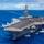 170612-N-BL637-144 A large US Navy aircraft carrier is underway in open blue ocean, viewed from an elevated aerial perspective. The flight deck is populated with numerous fixed-wing aircraft and personnel in colored jerseys performing flight deck operations. A second vessel is visible in the distant background.