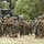 170614-N-PF515-013 A large group of military personnel in full combat gear are assembled in an open field area surrounded by military vehicles and wooded terrain. The soldiers are wearing MARPAT (Marine pattern) camouflage uniforms, suggesting these are U.S. Marines. The scene appears to be a staging or briefing area during a field exercise or operational deployment.
