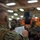 170614-N-GG458-047 A military briefing or muster is taking place in what appears to be a ship's interior bay or hangar deck. A service member in MARPAT digital camouflage uniform is reading from a document to a group of assembled personnel, several of whom have camouflage face paint applied. The setting and uniforms are consistent with U.S. Marine Corps or Navy personnel preparing for an operation.