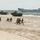 170614-N-PF515-010 A military amphibious assault exercise is underway on a sandy beach, with soldiers in combat gear running inland after disembarking from Amphibious Assault Vehicles (AAVs). Two AAV-7 tracked amphibious vehicles are visible on the shoreline, while at least two naval vessels, including what appears to be a landing ship bearing hull number 823, are positioned offshore. The scene depicts a coordinated beach landing operation consistent with NATO or allied military exercises.