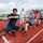Sterett-Dewey Surface Action Group Deployment A group of people are engaged in a tug-of-war competition on a red athletic track. Several participants are wearing blue-and-white striped shirts, while one person in the foreground wears a dark navy shirt. A uniformed official in white stands in the background observing, with a building displaying Chinese characters visible behind them.