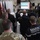 170614-N-JI086-354 A multinational military briefing or ceremony is taking place aboard a U.S. Navy vessel, identifiable by the ship's deck markings (LPD 2x). Personnel from multiple nations are gathered facing a speaker at a podium, with a projection screen displaying colored slides. Attendees wear a variety of military uniforms including U.S. Army multicam, French Marine Nationale, naval dress uniforms, and other allied nation attire.