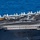 170612-N-YA681-007 An aerial view of a U.S. Navy aircraft carrier flight deck showing active flight operations at sea. Multiple fixed-wing aircraft are staged across the deck while at least one aircraft appears to be in the process of a catapult launch, evidenced by steam/vapor trails on the runway. Deck crew in color-coded jerseys are visible performing various flight deck roles.