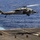 170612-N-LK571-423 A military helicopter, identified as an MH-60 Seahawk (tail number 13), hovers over the deck of a naval vessel at sea. A rope or fast-rope line descends from the helicopter's cabin to the flight deck below, where four personnel in combat gear are crouched in position. This appears to be a fast-rope insertion/extraction (FRIES) or helicopter rope suspension technique (HRST) training exercise.