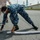 170615-N-YD204-013 A U.S. Navy sailor in blue digital camouflage (NWU Type I) uniform is bent over deploying an absorbent boom or containment sock on an asphalt surface, likely during a spill response drill or environmental training exercise. A second individual in tan/khaki coveralls stands nearby holding what appears to be a radio or tool. The setting appears to be a military installation with industrial infrastructure visible in the background.