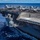 170612-N-BL637-194 An aerial-angle photograph of a large US Navy aircraft carrier underway at sea, showing the flight deck populated with multiple military jet aircraft. Personnel in colored jerseys are visible on the deck performing flight operations duties. A second vessel is faintly visible on the horizon in the background.