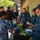 NMCSD NEC Fair A group of U.S. Navy personnel in blue digital camouflage uniforms are gathered around an outdoor display table at what appears to be a military information or demonstration event. The table contains security and law enforcement equipment including caution tape, a Garrett metal detector wand, a ballistic helmet, and candy. The setting appears to be a courtyard or walkway of a building with additional military personnel visible in the background.