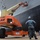 USS Blue Ridge Sailors give the ship a new coat of paint A U.S. Navy sailor in digital camouflage uniform and hard hat observes maintenance work being performed on the hull of a large naval vessel in a dry dock. A JLG Ultra Boom articulating boom lift (model 1200SJP) with an orange and yellow color scheme is extended toward the ship's hull, with a worker in the elevated platform basket. The equipment bears a U.S. Government license plate numbered N54-09749.