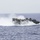 170602-N-FM530-182 A U.S. Navy Landing Craft Air Cushion (LCAC) is traveling at high speed across open ocean waters. The hovercraft is generating a large spray wake and visible air cushion exhaust as it skims the surface. The vessel displays 'NAVY' markings and an American flag on its hull.