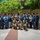NMCSD NEC Fair A large group of approximately 30 military personnel pose for a formal group photograph outdoors in a courtyard or plaza area. The group includes members wearing multiple uniform types, including Navy Working Uniform (NWU) blue digital camouflage, tan/khaki service uniforms, and woodland/desert camouflage patterns. The setting appears to be a military installation or government facility during daytime with bright sunlight.