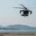 170615-N-SD711-0029 A military helicopter, consistent with a UH-60 Black Hawk design, is captured in flight over a coastal airfield or helipad. The aircraft appears to be either landing or taking off, with rotor blades in motion and a landing light illuminated. Mountains and a body of water are visible in the background, suggesting a coastal or island military installation.