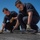 170613-N-VN584-210 Two personnel wearing dark navy coveralls and work gloves are crouched on what appears to be a ship's flight deck, performing maintenance work on a metal track or rail component. They are working collaboratively on a low-profile metal fixture secured to the deck surface. A power drill is visible nearby, and additional personnel can be seen in the background.