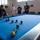 Sterett-Dewey Surface Action Group Deployment A group of people are gathered around a blue-felt pool/billiards table in what appears to be a recreational facility. A young blonde man is actively taking a shot while others, including a person in a white military-style uniform, watch. The setting suggests a cross-cultural social or diplomatic gathering.